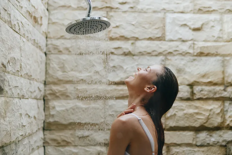woman enjoying outdoor shower with stone wall background, wet hair, relaxed mood, natural light, healthy skin, summer wellness concept