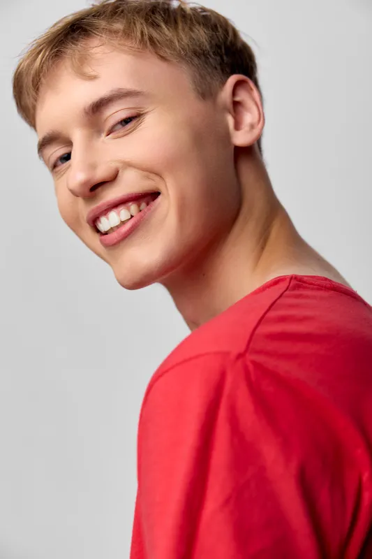 Smiling man wearing a red shirt stands against a neutral background, conveying confidence and approachable energy. Ideal for lifestyle, fashion, or wellness topics featuring positivity and friendly