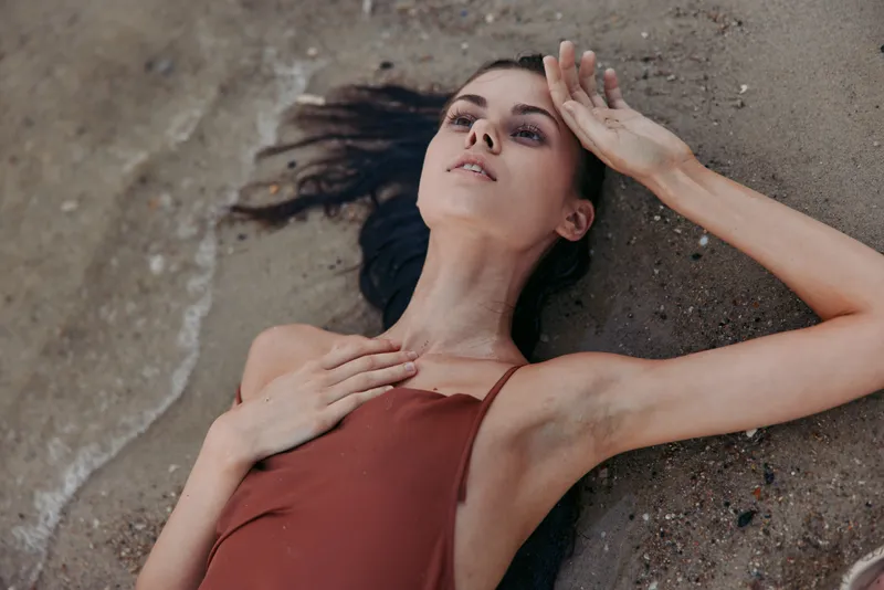 Relaxed woman lying on the sand near the water, wearing a brown swimsuit with calm expression in a natural beach setting.