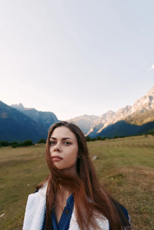 woman portrait in mountains meadow, nature landscape outdoors scene with young traveler looking into camera, long hair and thoughtful expression in wide alpine field at golden hour