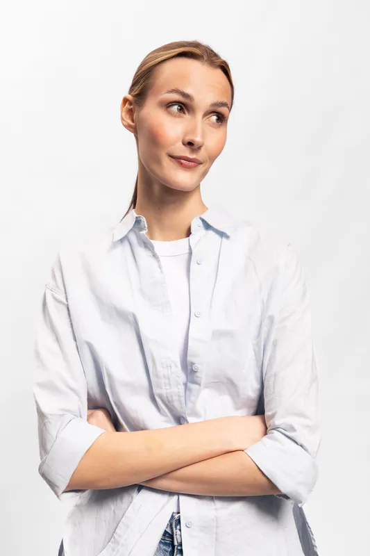 Young woman in casual white shirt stands with crossed arms looking sideways. Portrait on white background with natural light. Concept for confidence, contemplation, and casual fashion.
