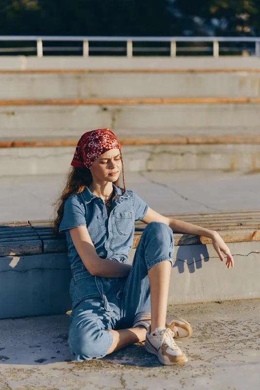Lifestyle woman in modern boho-western denim street style wearing a red bandana, sitting relaxed on concrete steps with warm film color tones and natural sunlight.