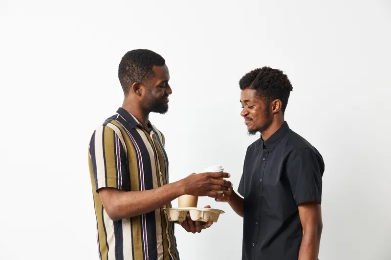 Two smiling black men sharing a coffee moment, dressed casually in stylish attire, creating a warm and friendly atmosphere together.