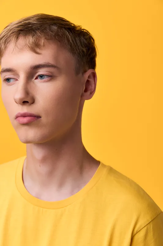 Closeup of a calm contemplative man wearing a yellow shirt against a solid yellow backdrop, minimal studio lighting, clean composition, soft shadows, thoughtful gaze, fashion study