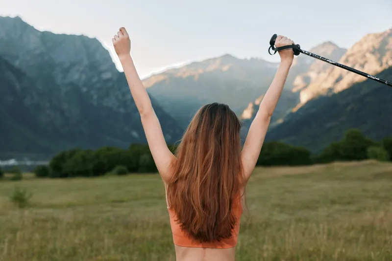 A person stands in a grassy field with arms raised high, gripping a trekking pole as mountains rise behind under a clear sky, conveying achievement and outdoor adventure.