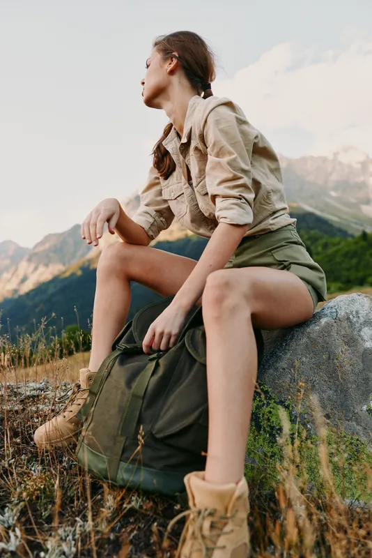 Young woman in casual outfit sitting on a rock with backpack in mountainous landscape. Outdoor lifestyle, adventure, hiking, nature exploration concept.