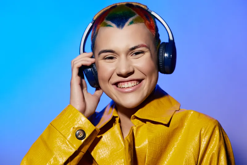 Smiling woman with colorful hair wearing headphones and a bright yellow jacket, enjoying music against a vibrant blue background.