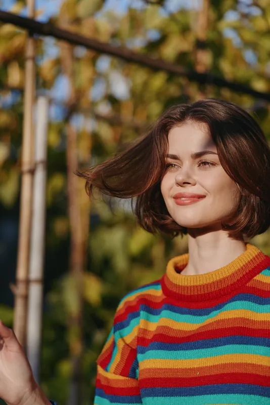 Young woman with short brown hair smiling in a colorful striped sweater outdoors in a sunny garden, touching hair with relaxed confident look, lifestyle portrait for fashion or travel.