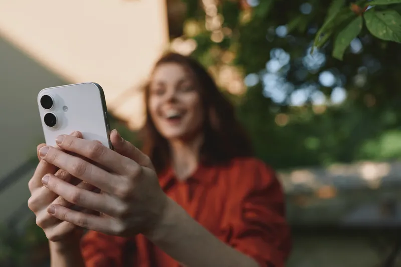 One smiling woman outdoors selfies with a white phone, wearing a red shirt, warm sunlight and green foliage in the background, casual pose and bright mood