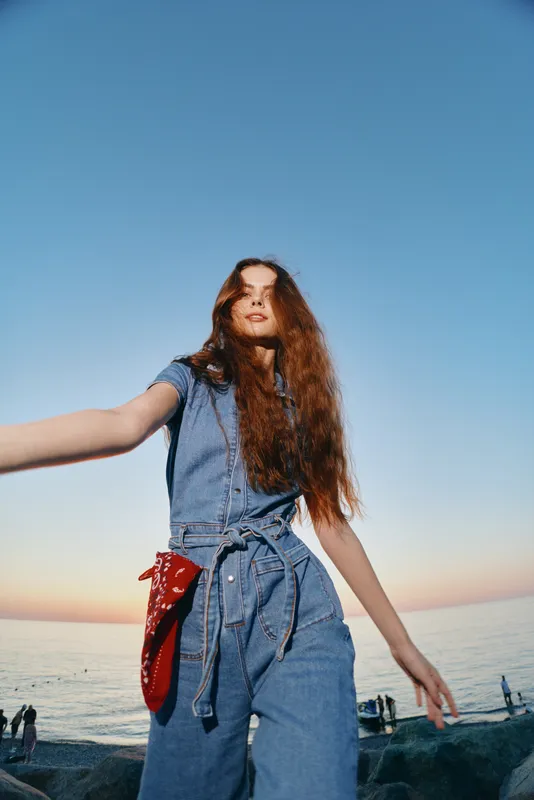 Lifestyle woman in modern boho-western denim street style with long hair by the sea during golden hour, film color enhancing casual youthful vibe and relaxed pose.