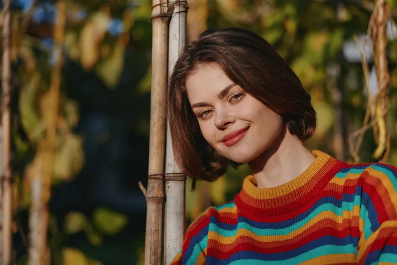 young woman in her early 20s wearing a colorful striped sweater, smiling and leaning on bamboo pole with short brown hair in a sunny outdoor garden for lifestyle fashion.
