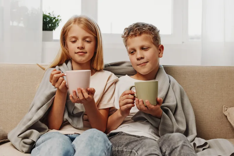 Two cozy children wrapped in grey blanket sit on beige sofa holding warm mugs. Soft natural light fills bright room suggesting comfort, relaxation, and peaceful family moments at home.
