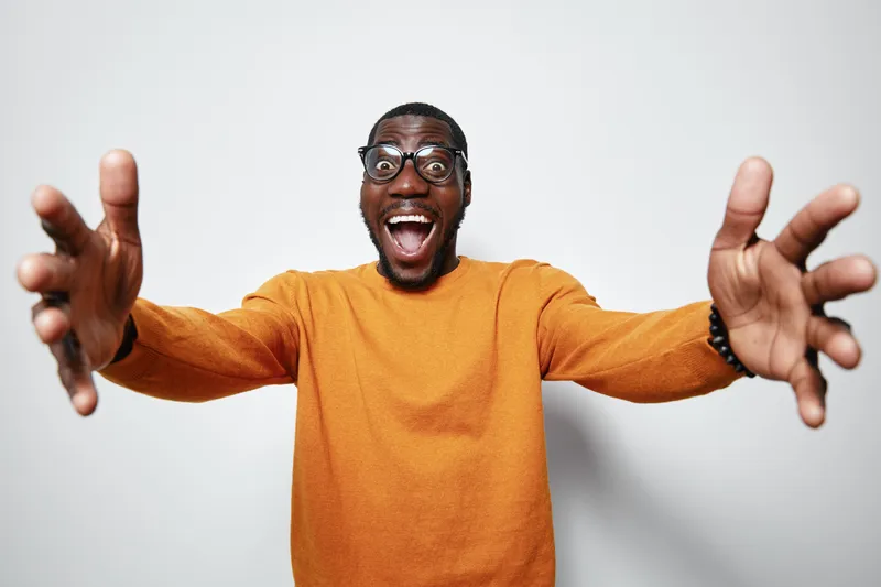 African man with dark skin and glasses wearing orange sweater smiling widely with open arms for hug or welcome on white background. Excited, happy and friendly expression.