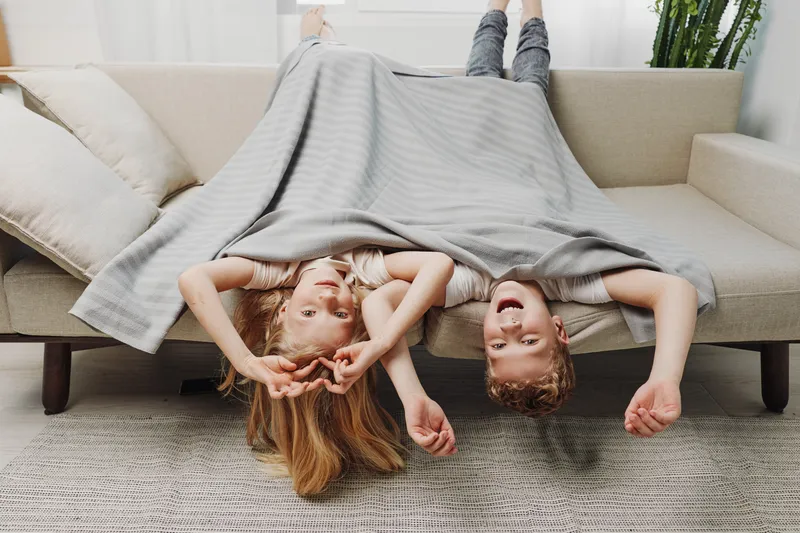 Happy children lying upside down on sofa covered with blanket, playing and smiling in cozy living room. Concept of family fun, childhood joy and indoor relaxation moments.