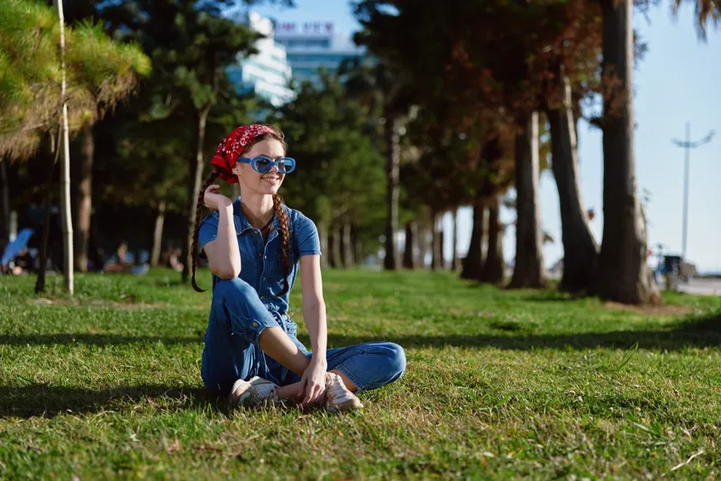 Lifestyle woman in modern boho-western denim street style sitting on grass outdoors wearing sunglasses and red bandana under bright sunlight film color.