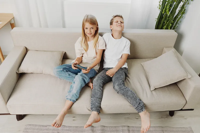 Two children relax barefoot on beige sofa in casual clothes, playing video games in bright living room with modern decor and natural light.
