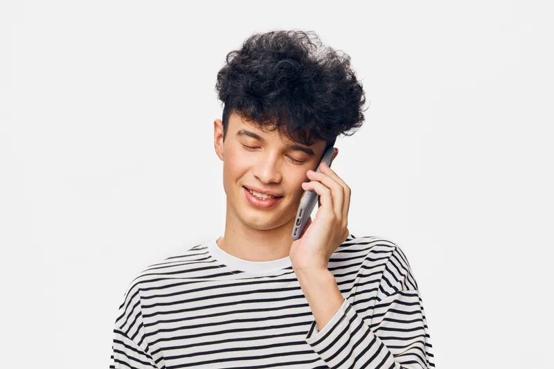 young man smiling while talking on smartphone, striped shirt, curly hair, casual look, isolated on white background, communication concept