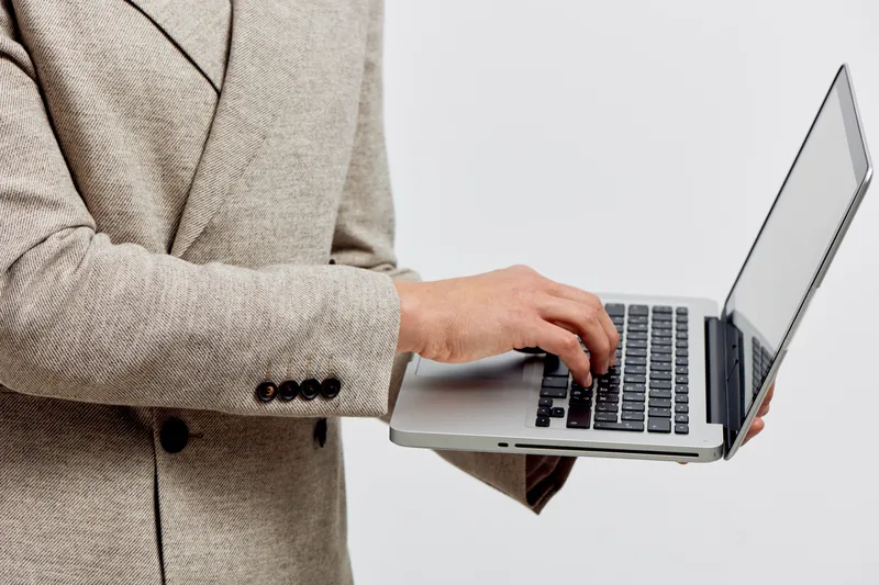 Professional young woman in beige blazer using silver laptop computer, standing against plain white background, close-up of hands and torso, online work and technology concept, modern office style.