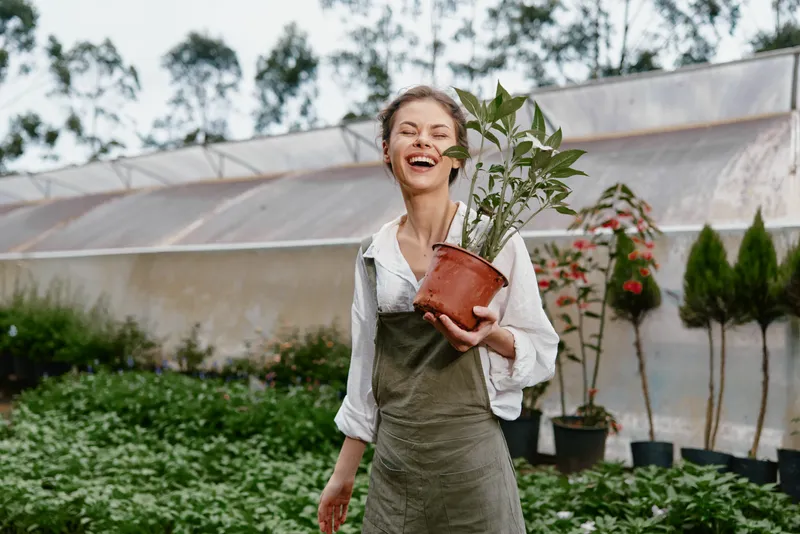 Young woman in an apron joyfully holding a potted plant in a greenhouse, surrounded by lush greenery and colorful flowers, embodying a love for gardening