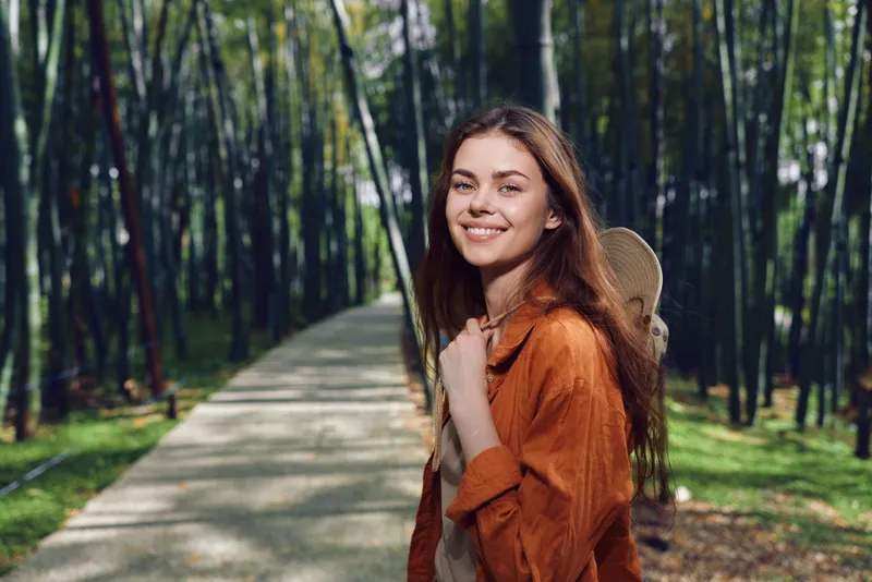 Woman smiling with skateboard and backpack on a park path through bamboo grove, casual youth portrait in nature, outdoor leisure and relaxed lifestyle moment.
