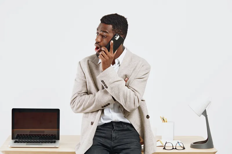 Professional young African American man wearing beige blazer and white shirt talking on mobile phone while standing at desk with laptop, eyeglasses, and lamp against white background. Business lifesty