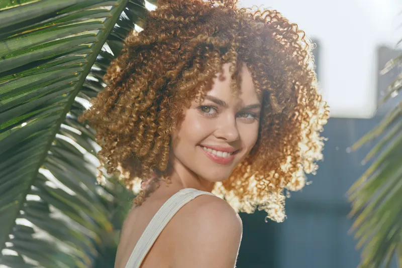 smiling woman with curly hair outdoors, natural light, tropical leaves background, youthful, happy, sunlight, warm mood