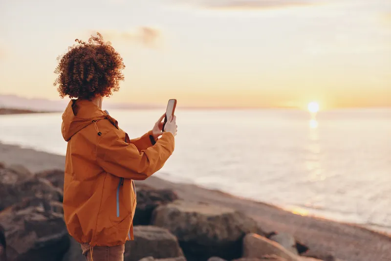 Curly hair child in orange jacket standing on beach at sunset, holding smartphone to take photo, ocean horizon behind, lifestyle travel moment, warm golden light, peaceful mood