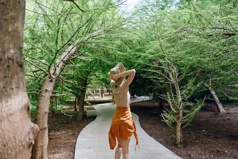 Woman walking on gravel path in forest wearing straw hat and orange skirt, back view. Leisurely summer stroll among green trees, natural light and peaceful nature scene outdoors.