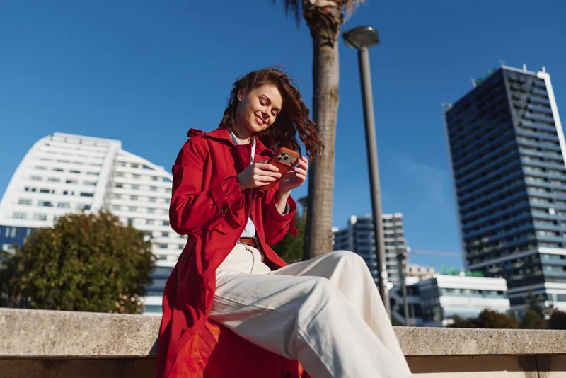 Young woman in red coat sitting outdoors using smartphone, smiling, sunny day, modern urban background, casual outfit, relaxed mood