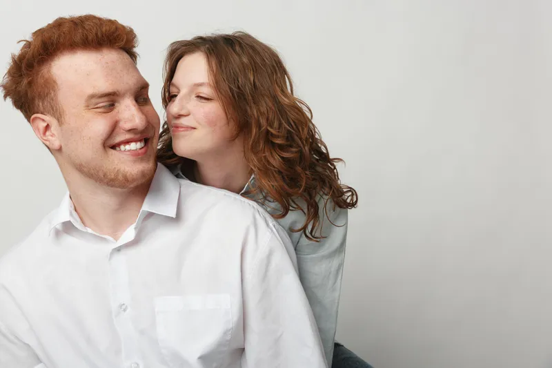 Smiling young couple with red hair and curly hair in casual white shirts share a tender moment on light background. Concept of love, affection, and happy relationship.