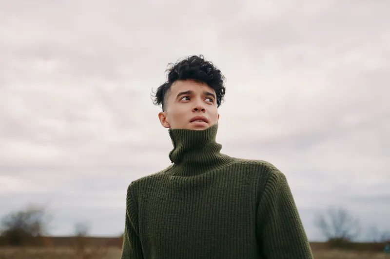 A candid portrait of a young man in a dark green turtleneck sweater, conveying authenticity and credibility through natural expression and a calm, thoughtful gaze against a muted sky.