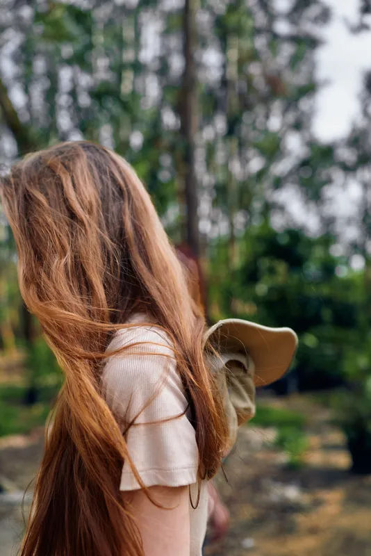 Woman with hair cascading long over her back, holding a hat behind her in a forest setting. Natural light, casual clothing, outdoor portrait capturing texture of hair and peaceful nature mood.