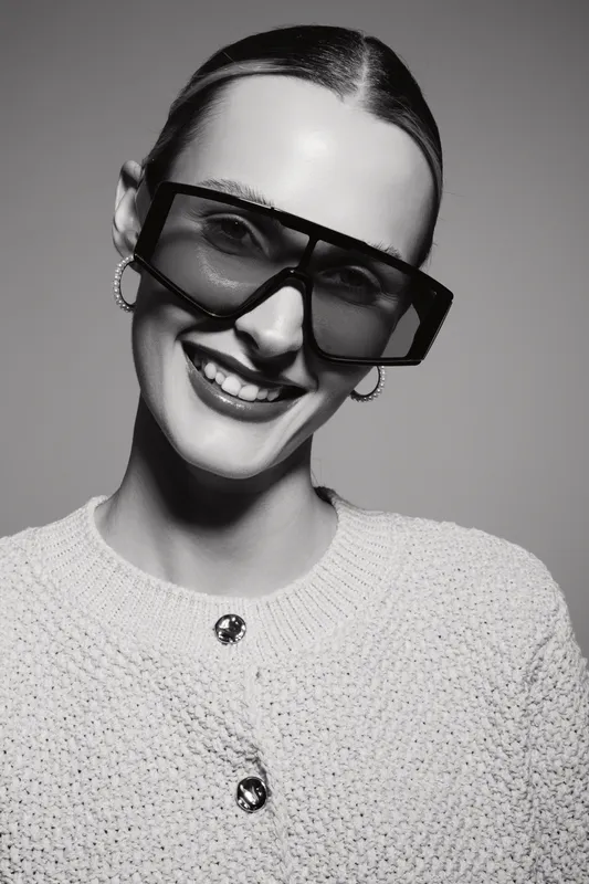 Smiling white woman with fair skin wearing large sunglasses and knit sweater in black and white studio portrait.