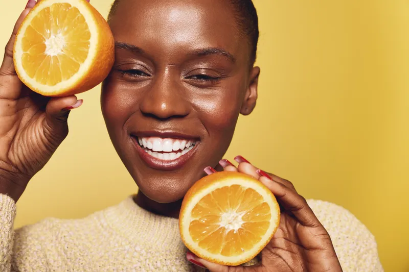Happy African woman with dark skin holds two orange halves near face, smiling with eyes closed. Yellow background and cozy sweater create warm, fresh, and healthy lifestyle concept.
