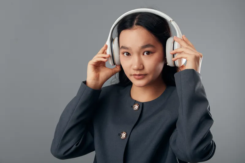 Young woman with headphones on gray background wearing dark jacket adjusting headset for music or audio listening, portrait of female with modern tech gadget and casual style.