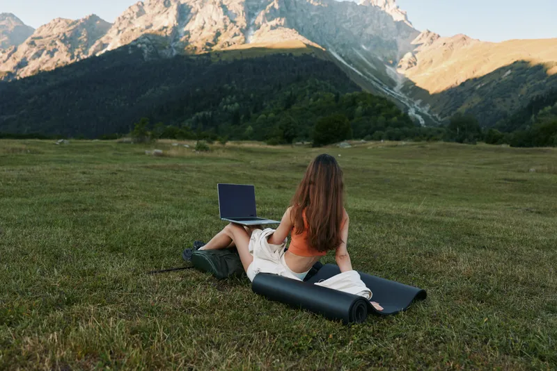 A woman sits on a grassy field with a laptop in front of a mountain landscape. She appears focused and relaxed, creating a calm, productive outdoor work vibe.