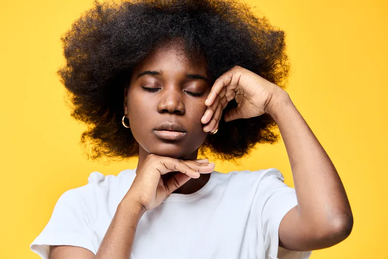 Young Black woman with natural hair in a white shirt, posing gracefully against a vibrant yellow background, conveying a sense of peace and tranquility