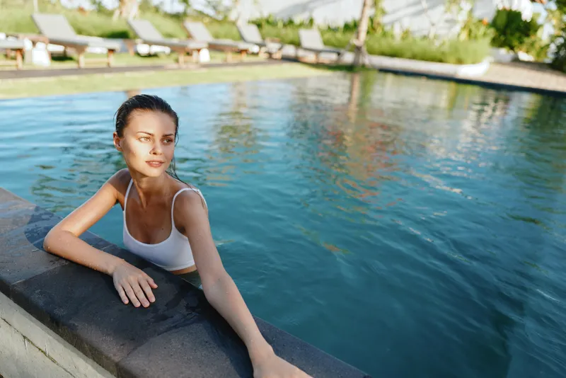 A pool, woman, swimming, relaxation, outdoor, sunny, water, leisure, lifestyle, summer scene shows a female enjoying the pool edge with a calm expression and casual attire in a bright resort setting