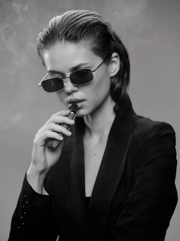 Young woman smoking electronic cigarette in black and white minimalistic style with sunglasses and dark clothing.