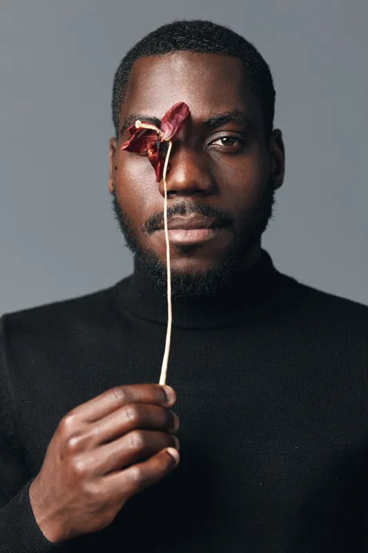African man with dark skin holding dried flower in front of face in creative conceptual visual style. Portrait with black turtleneck and neutral background for artistic expression.
