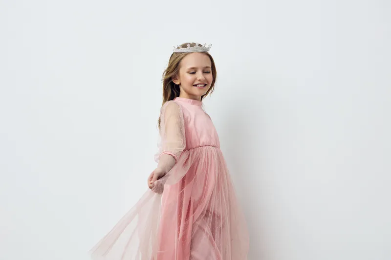 Happy young girl in a pink dress with a crown, twirling joyfully against a light backdrop, embodying innocence and celebration in a stylish, whimsical way
