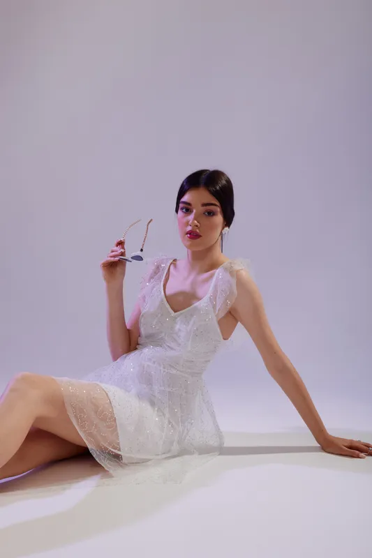 Young woman in a white lace dress poses seated on the floor holding sunglasses. Soft lighting highlights her beauty and fashion style against a plain background. Concept of elegance and style.