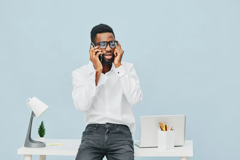 Confused young man with beard and glasses wearing white shirt and jeans, holding cellphone to his ears, standing near office desk with laptop and stationery, on plain light blue background, studio sho