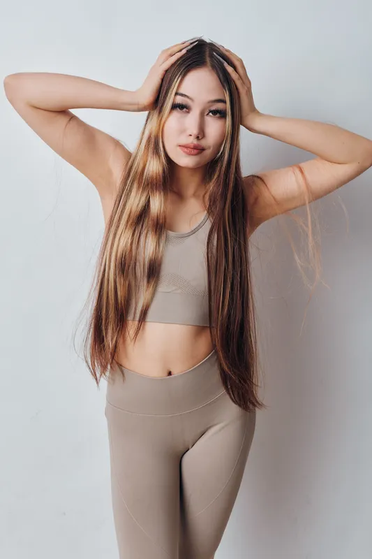 Young woman with long straight hair wearing beige sportswear posing with hands on head against light background for beauty and fashion portrait concept.