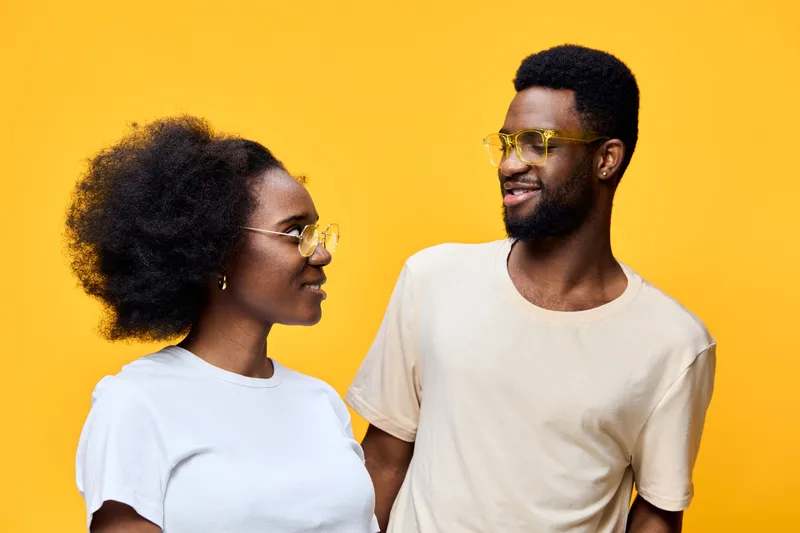 Young multi ethnic couple smiling joyfully against a vibrant yellow background, wearing stylish summer outfits and trendy sunglasses, conveying love and happiness