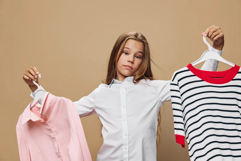 Girl holding two different shirts on hangers for choice and comparison. Young female with casual and formal clothes in studio for fashion decision or style selection.