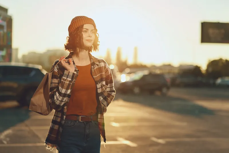 Woman urban golden candid lifestyle authenticity beanie confident portrait of a young shopper walking through a sunlit parking lot at golden hour, relaxed, mindful living and emotional storytelling.