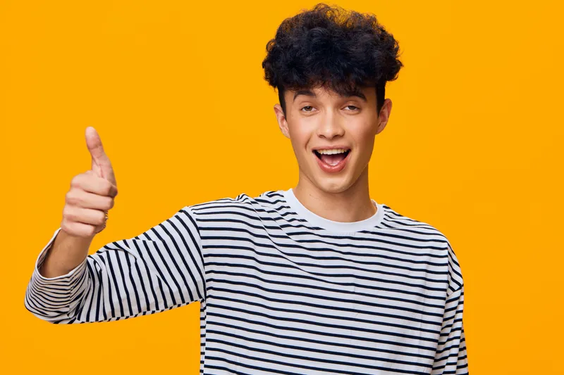 young man showing thumbs up wearing striped shirt with curly hair on bright orange background, expressing positivity and enthusiasm, casual style