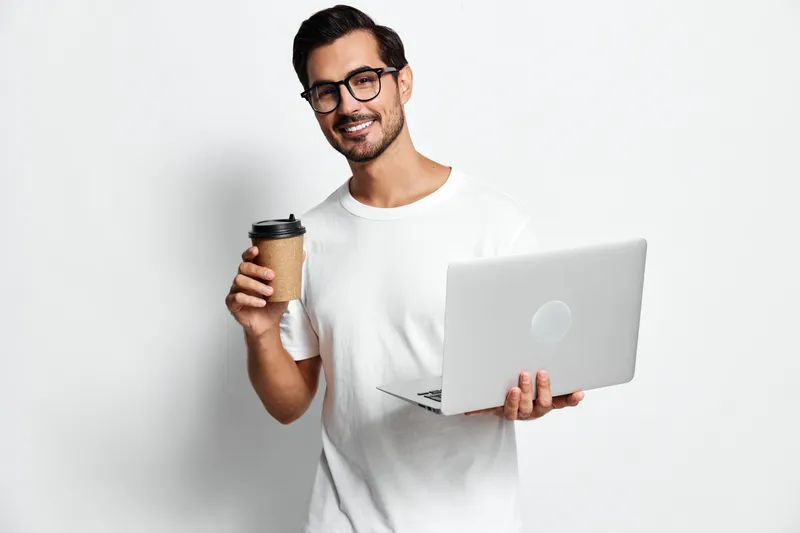 Smiling man holding coffee cup and laptop wearing casual t-shirt, looking at camera with cheerful expression, isolated on plain light background. People lifestyle concept.