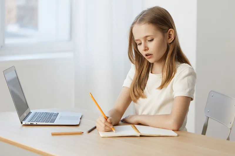 Young girl studying at a desk, focused on her notebook, with a laptop in a bright, minimalistic setting, embodying learning and concentration